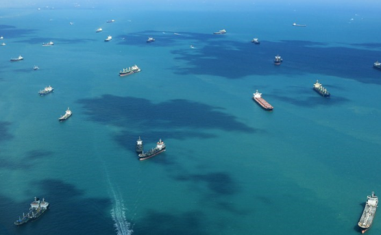 Aerial view of a busy maritime traffic lane showcasing numerous ships navigating through turquoise waters, some emitting plumes from bunker fuel.