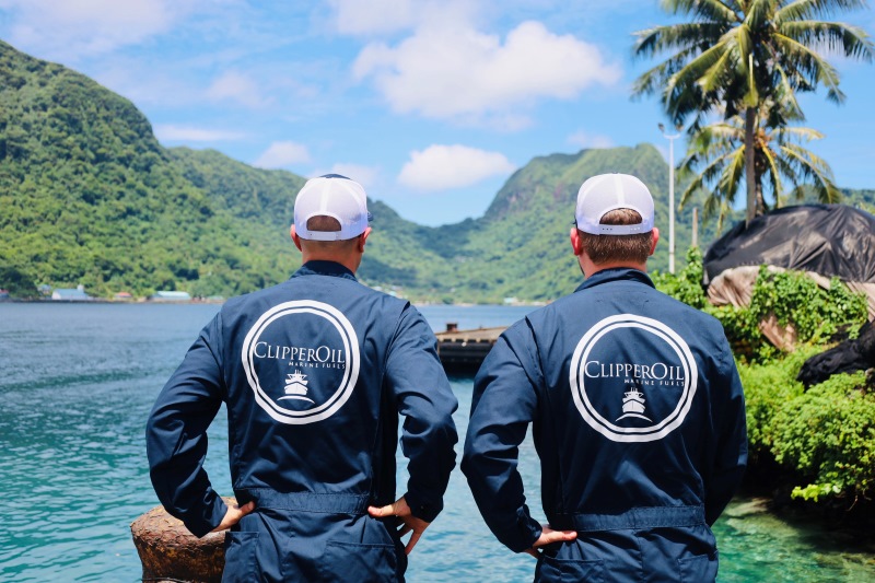 Two individuals in matching uniforms with "clipper oil" logos on their backs, overlooking a tranquil bay with lush green mountains in the background.