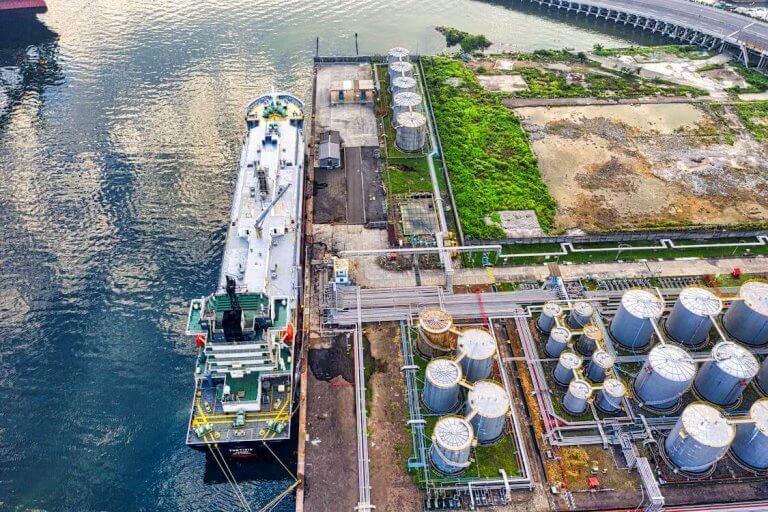 Aerial view of an industrial port showing a large ship docked alongside a facility with multiple storage tanks. The scene includes water on the left, various pipelines, and a mix of greenery and cleared land in the background.