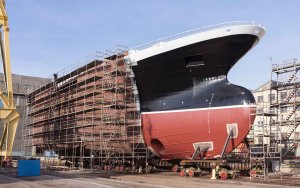 A large ship under construction in a shipyard is surrounded by scaffolding and industrial equipment. The front section of the ship is painted black and red, while the rest of the hull appears unfinished with a rust-colored surface. The ground is cluttered with construction materials.