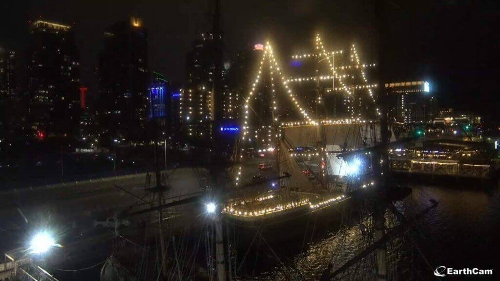 A beautifully illuminated tall ship with string lights is docked at the waterfront, captured via a San Diego webcam, against the night backdrop of a city skyline. The image showcases the serene and festive harbor atmosphere with bright reflections shimmering on the water.